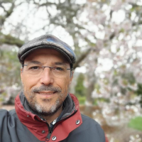 Fotografia de l'autor del blog. Primer pla lleugerament descentrat d'un home blanc de mitjana edat, somriu, té una barba curta canosa, duu ulleres, una gorra plana de llana en el cap, vesteix una jaqueta esportiva roja i grisa de la qual només es veu la part de dalt. Al fons s'aprecia de forma desenfocada un arbre probablement un cirerer florit i un paisatge verd.